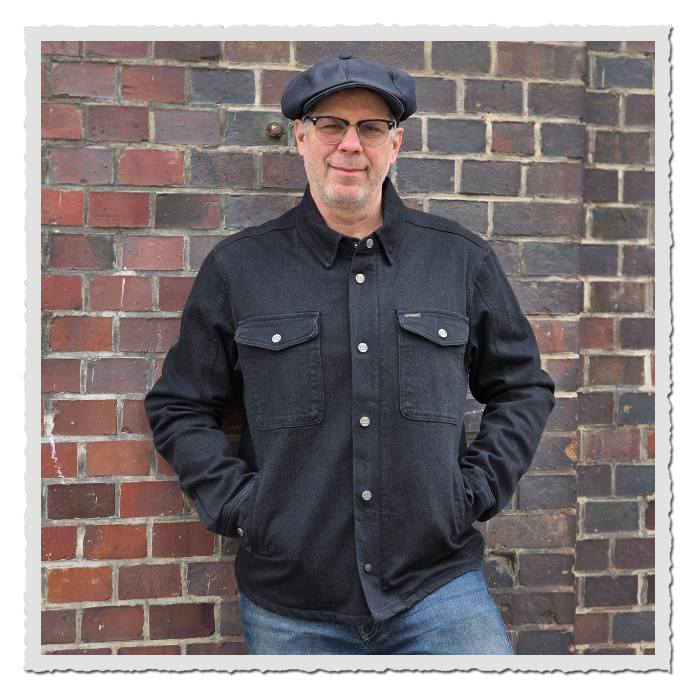 FroFrontalansicht einer Person im dunklen Rider Shirt, Brille und Mütze vor einer gemauerten Backsteinwand im Außenbereich.ntalansicht einer Person in dem dunklem Rider Shirt, Brille und Mütze vor einer gemauerten Backsteinwand im Außenbereich.